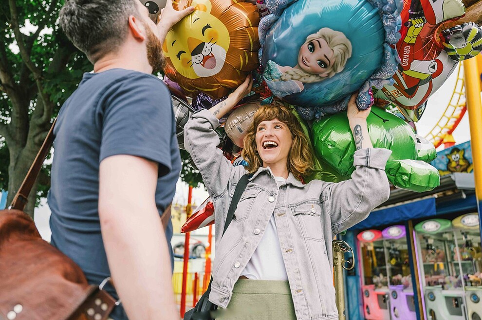 Wiener Prater: Eine Frau steht lachend unter einer Traube bunter Luftballons. Ihr gegenüber steht ein Mann. Förderungen, Beratungen, Events in der Wirtschaftsagentur Wien. 
