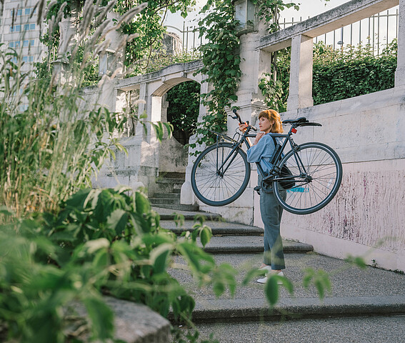 Nachhaltig Handeln: Förderungen, Beratungen, Workshops für Unternehmen. Eine Frau im Park trägt ein Fahrrad Stufen rauf