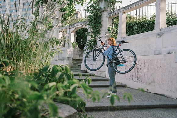 Nachhaltig Handeln: Förderungen, Beratungen, Workshops für Unternehmen. Eine Frau im Park trägt ein Fahrrad Stufen rauf