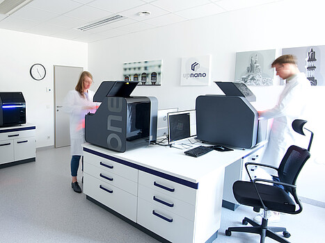 Two people in white lab coats work in a laboratory room with modern equipment, including a NanoOne technology device.