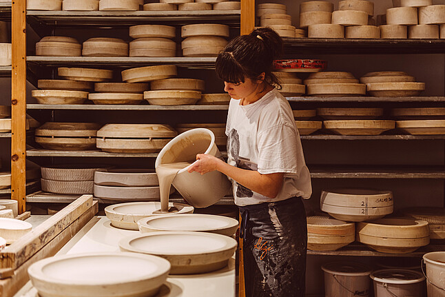 feinedinge: Eine Frau steht in einem Atelier voller Keramik und gießt flüssigen Gips in eine Form. 