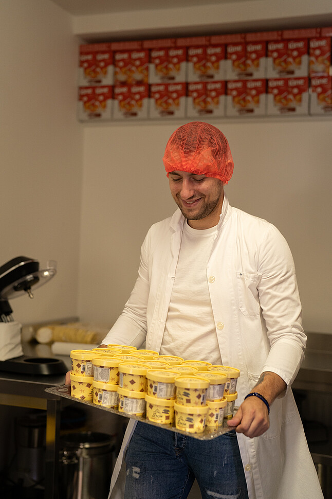 [Translate to English:] Dajan Srejic trägt ein Haarnetz und einen weißen Kittel und hält ein Tablett mit gelben Eisbechern. Im Hintergrund sind weitere Produkte in Regalen zu sehen.