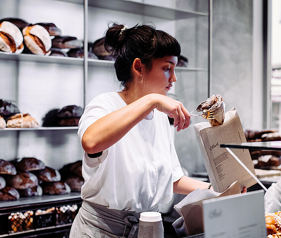 Graetzel beleben: Förderungen, Beratungen, Netzwerke für Unternehmen in Wien. Eine Frau in einer Bäckerei steckt ein Stück Gebäck in ein Verkaufssackerl.