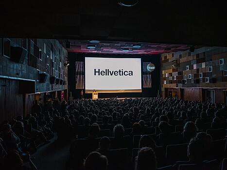 Menschen sitzen in einem abgedunkelten Kinosaal, auf der Leinwand steht das Wort Hellvetica