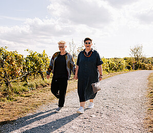 Bistra und Christa: Zwei Frauen gehen Hand in Hand auf einem Schotterweg in den Weinbergen spazieren