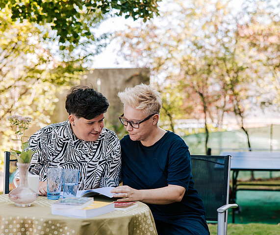 Two women sitting at a table, reading a book together.