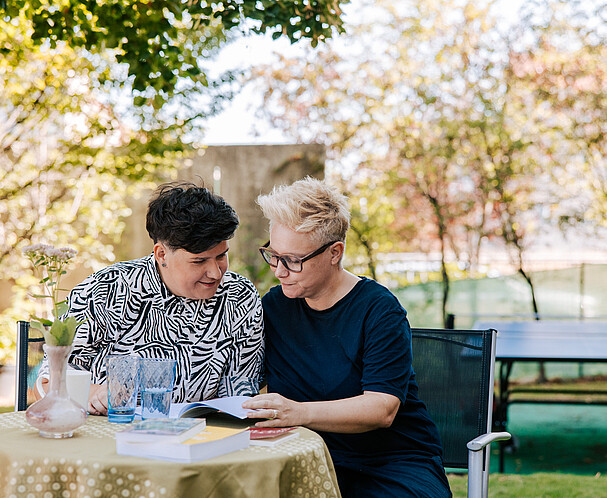 Two women sitting at a table, reading a book together.