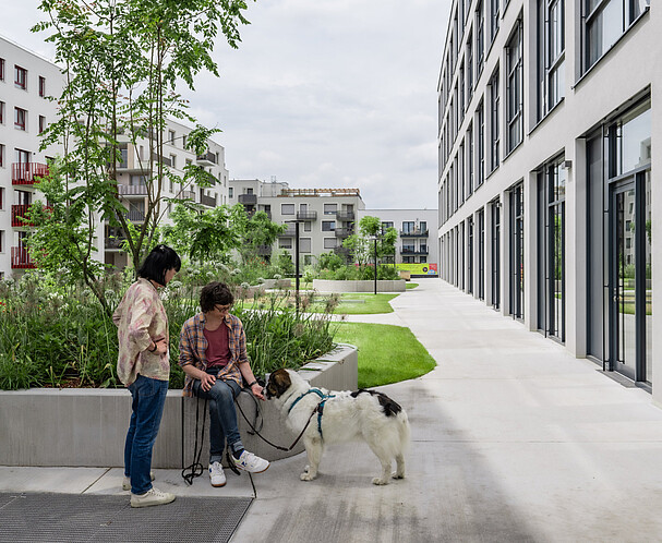 Eine stehende und eine sitzende Frau mit einem Hund befinden sich auf einer Grünfläche vor dem Gewerbehof Seestadt 