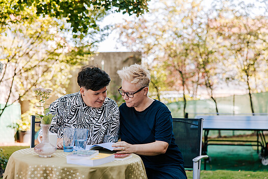 Bistra und Christra: Zwei Frauen sitzen im Garten an einem Tisch und sehen sich gemeinsam ein Buch an.