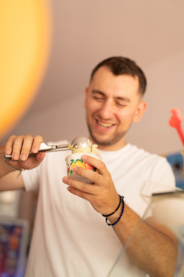 [Translate to English:] Dajan Srejic, Gründer von Sreja Ice, steht hinter der Theke und lächelt, während er eine Kugel Eis in einen Becher füllt. Er trägt ein weißes T-Shirt und ein Armband, das Eis wird sorgfältig mit einem Eiskugler portioniert.